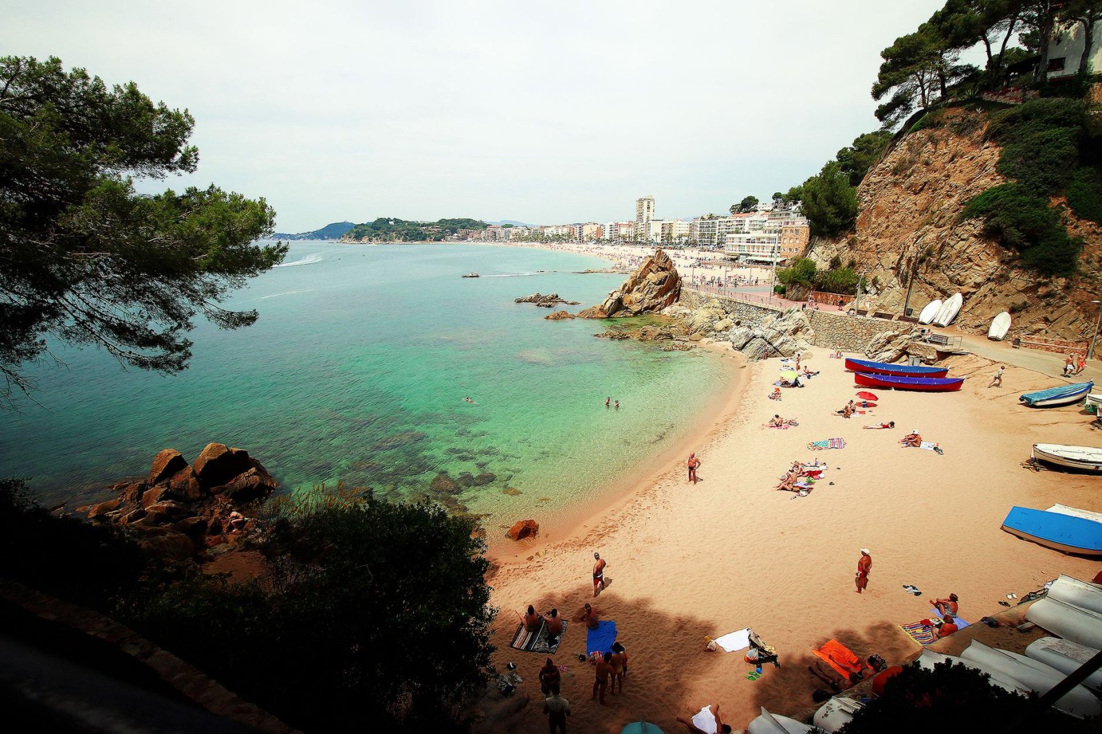 Playa mediterr&aacute;nea id&iacute;lica con aguas turquesas, arena y gente disfrutando del sol.